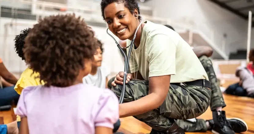 Smiling military service member using a stethoscope to check a young child’s heartbeat, demonstrating community healthcare and support.